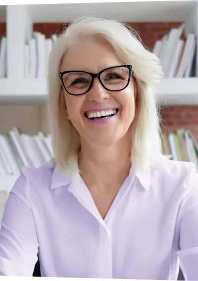 Mulher loira de olhos castanhos, camisa lilás, óculos, sorrindo em frente a uma estante de livros.