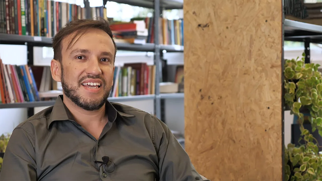 Rafael Matsudo, homem branco, cabelos e olhos castanhos, de barda e veste terno cinza com camisa cinza em uma biblioteca escolar.